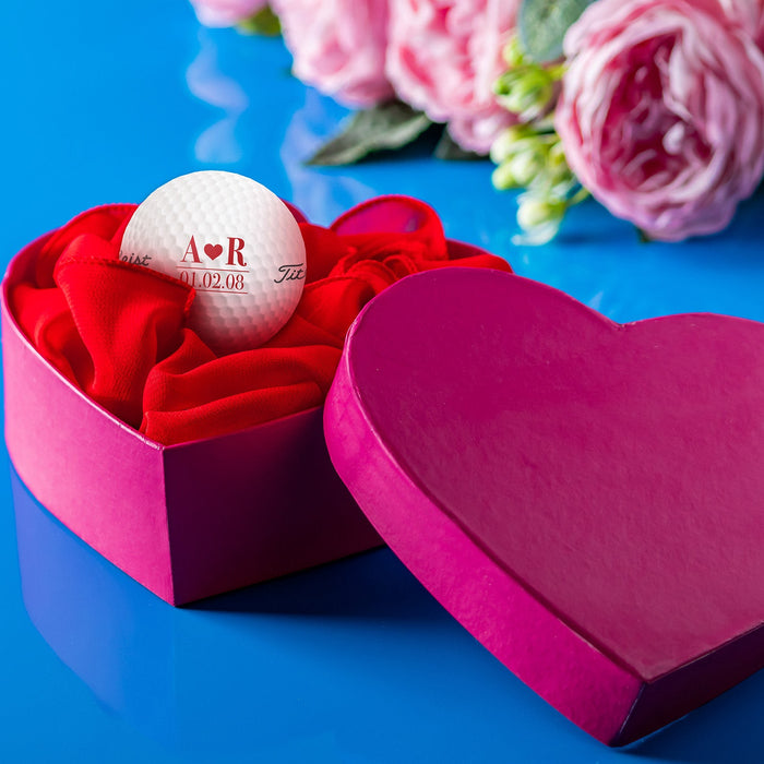 A pink heart shaped box sits open with a Valentine's Day golf ball inside. Both the box and a bouquet of pink roses sit on a blue surface.