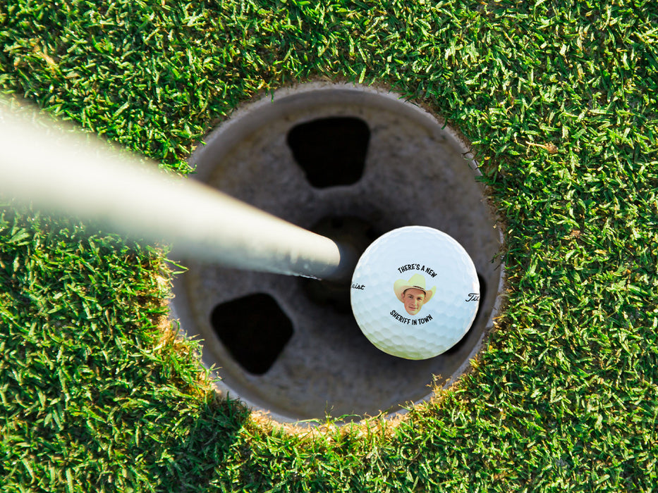 A golf ball is shown in a golf course hole. Ball features custom text and photo design.