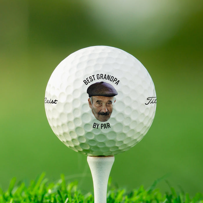 A golf ball is shown sitting on a golf tee with a grassy, green background behind it. Ball features custom printed text and photo.