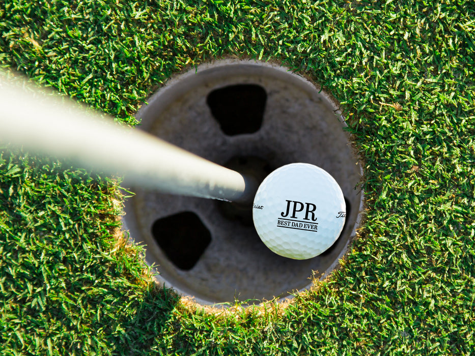 single white titleist golf ball with customizable personalized initial Best Dad Ever design in golf course hole next to pole surrounded by golf course grass