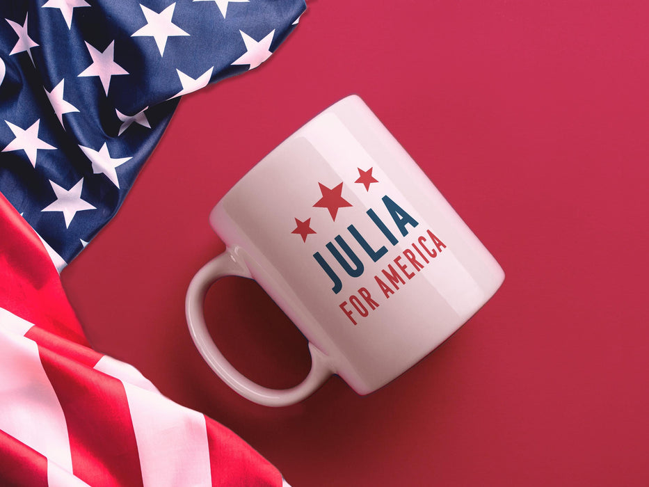 white mug with red white and blue American design with typography that says Julia for America with Red Stars sitting on a red surface next to an American Flag