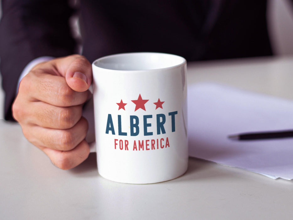 businessman sitting at desk with a white mug with red white and blue American design with typography that says Albert for America with Red Stars