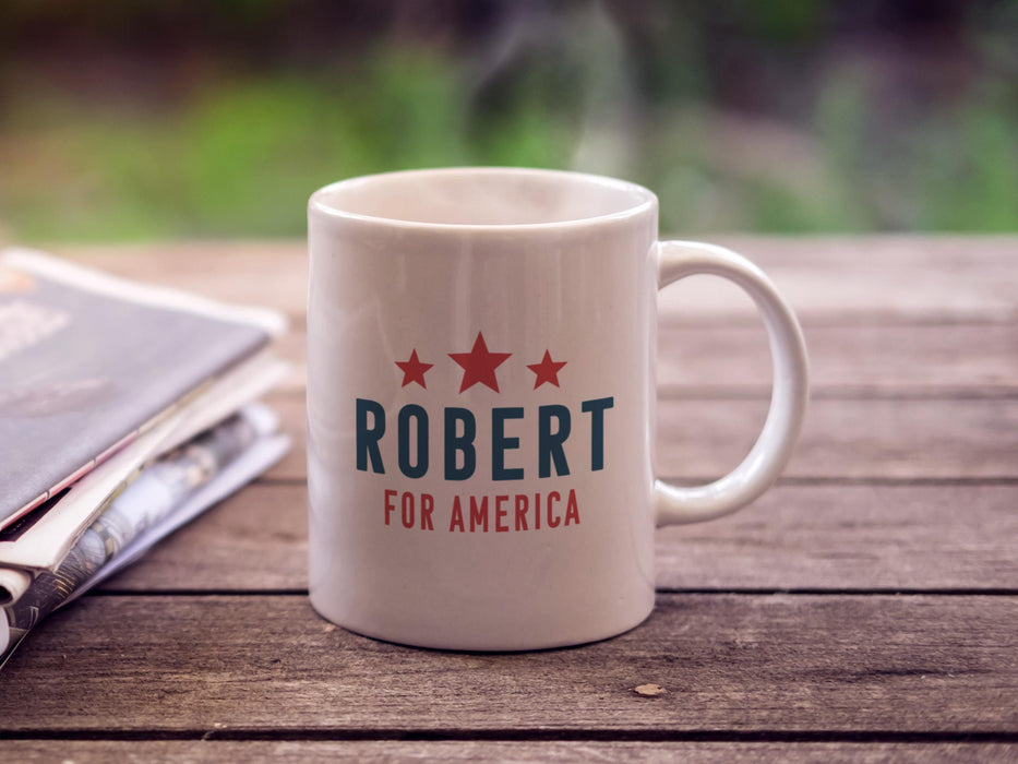 white mug with red white and blue American design with typography that says Robert for America with Red Stars sitting on a picnic table next to newspapers