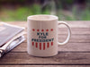 white mug with red white and blue American design with typography that says Kyle for America with Red Stars sitting on a picnic table next to newspapers