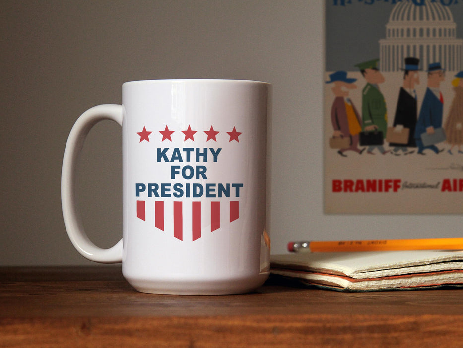 white mug with red white and blue American design with typography that says Kathy for America with Red Stars and Stripes sitting on a desk next to a notebook and pencil