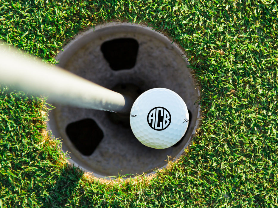 single white titleist golf ball with custom personalized black monogram printed design in golf course hole next to pole surrounded by golf course grass