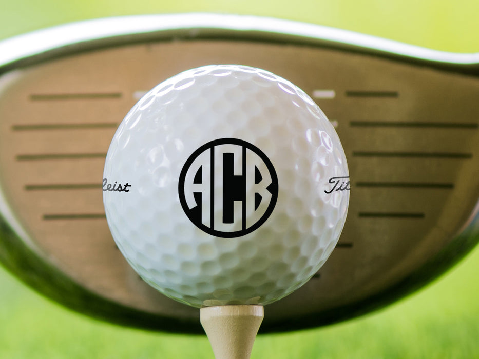 single white titleist golf ball with custom personalized black monogram printed design on top of beige golf tee with grass and golf club head in the background