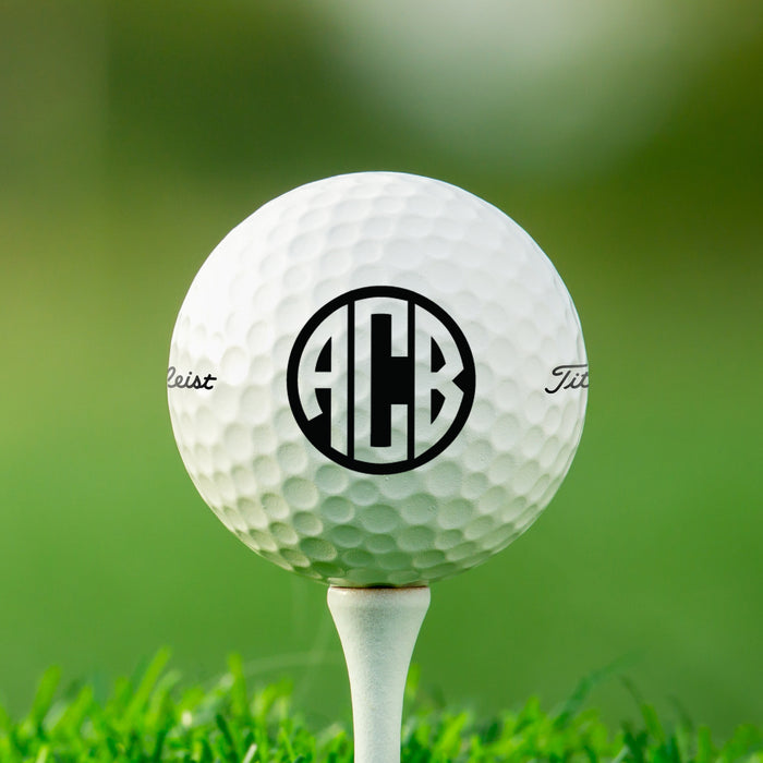 single white titleist golf ball on white golf tee with custom personalized black circle monogram ABC printed on it against grass golf field background