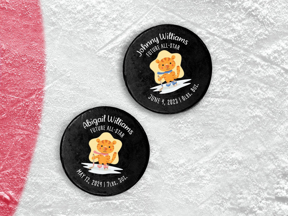 two hockey pucks on top of ice rink with future all star cartoon tiger designs with different names, birthdays, weights, blue and pink color options