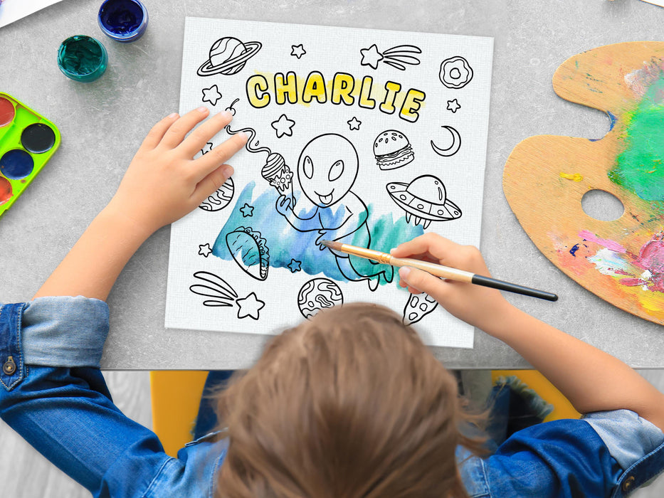 kid painting a canvas panel with a custom space alien adventure design printed on it, kid is using watercolors on grey table surrounded by painting supplies such as cups of paint, palettes, brushes, etc