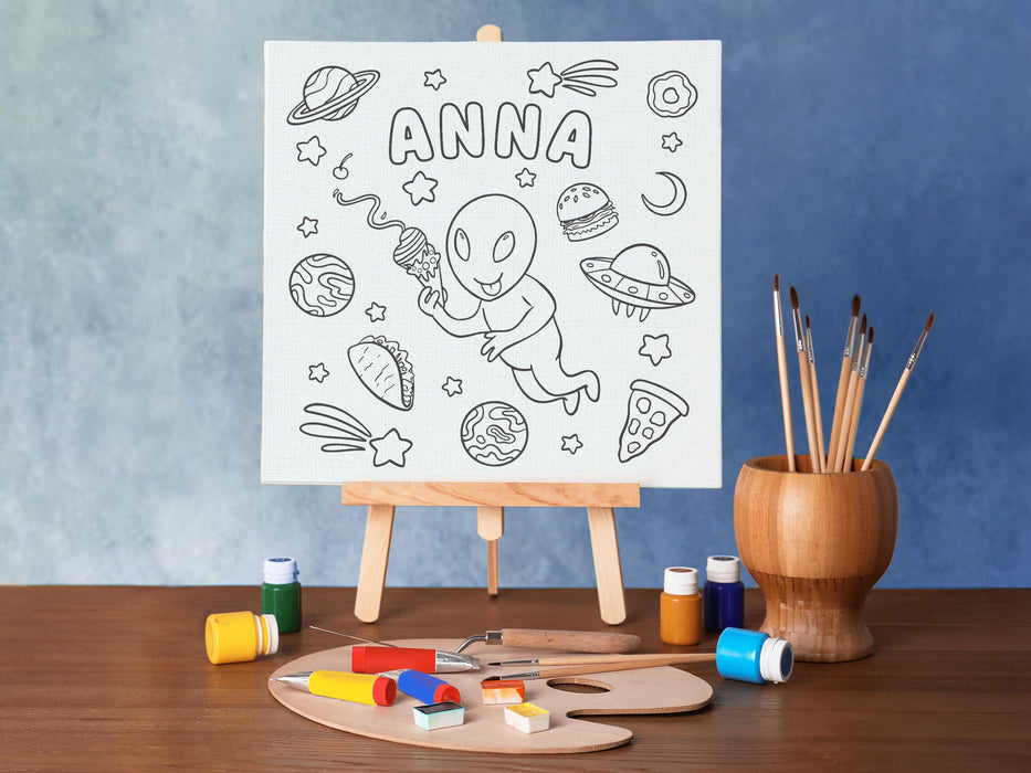 canvas panel with a custom space alien adventure design printed on it sitting on wooden easel, placed on a white wooden table surrounded by painting supplies such as paint brushes, tubes and paint palettes, palette knife, in front of a blue wall