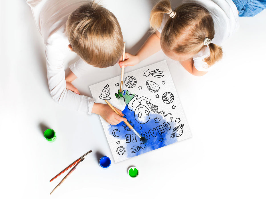 two kids on white floor painting a a custom space alien adventure design printed on it with blue and green paint as well as paint brushes