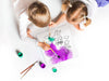 two kids on white floor painting a a custom caticorn design printed on it with teal and purple paint as well as paint brushes