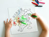 kid at white table coloring a canvas panel with a custom caticorn design printed on it using crayons with a pile of crayons right next to the canvas