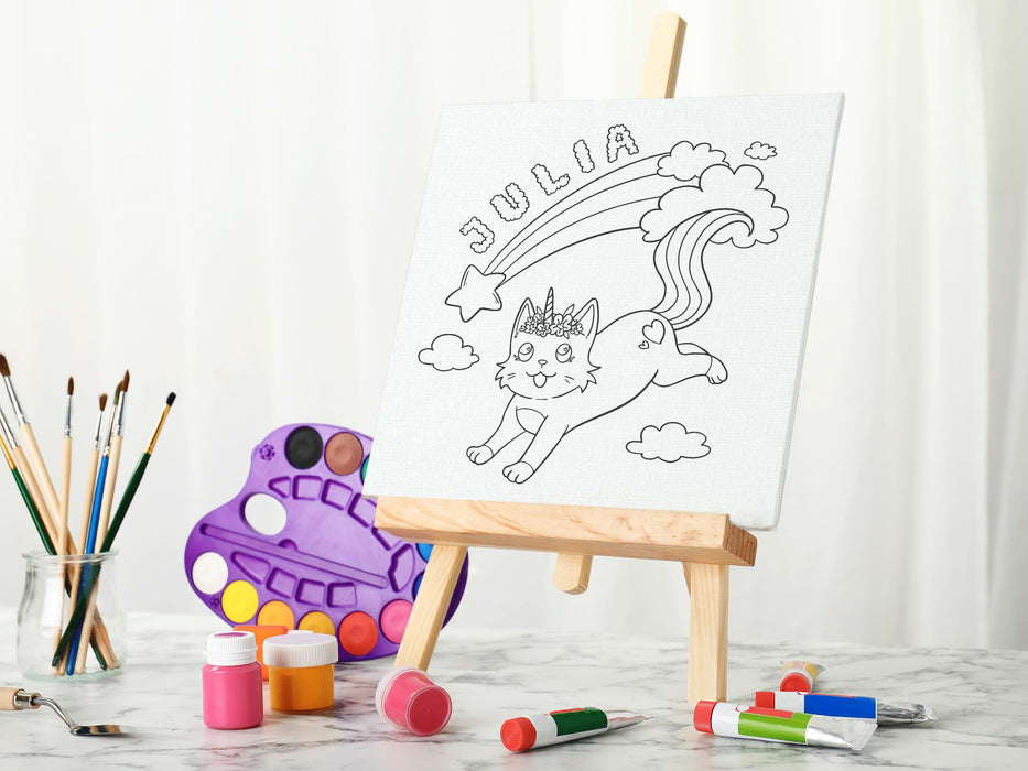 canvas panel with a custom cute caticorn design printed on it sitting on wooden easel, placed on a white marble wooden table surrounded by painting supplies such as paint brushes, tubes and paint palettes, palette knife, in front of a white curtain