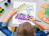 kid painting a canvas panel with a custom name caticorn design printed on it, kid is using watercolors on grey table surrounded by painting supplies such as cups of paint, palettes, brushes, etc