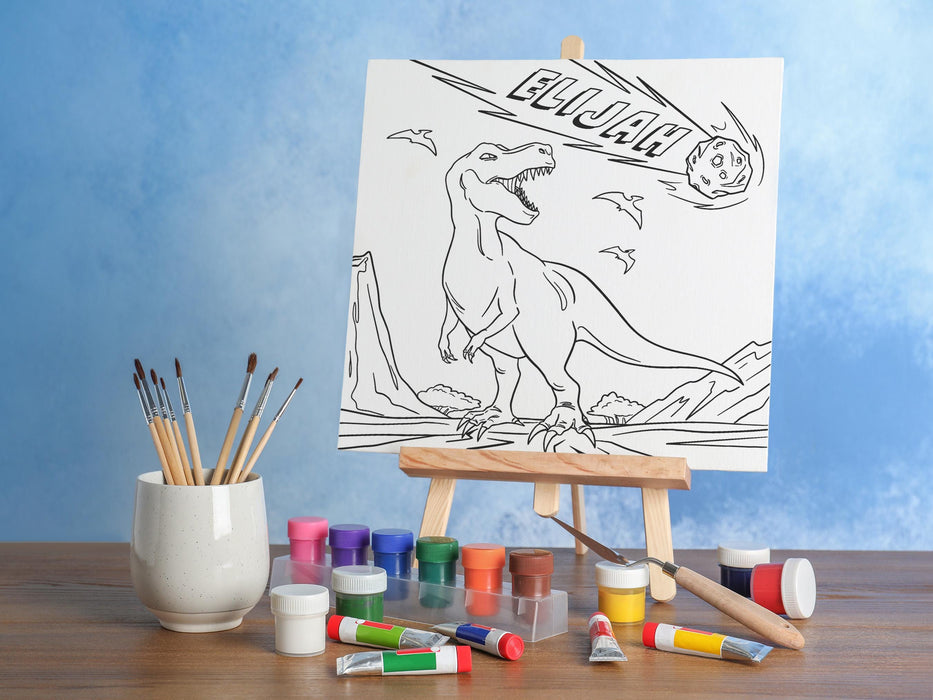 canvas panel with a custom name dinosaur t-rex design printed on it sitting on wooden easel, placed on a brown wooden table surrounded by painting supplies such as paint brushes, tubes and paint palettes, palette knife, in front of a blue wall