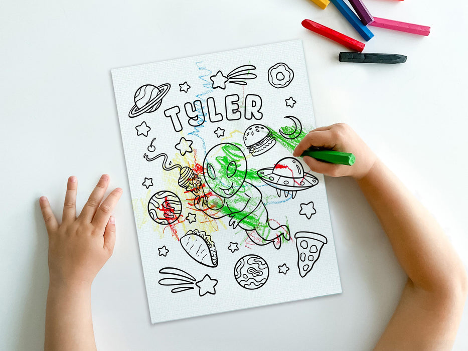 kid at white table coloring a canvas panel with a custom space alien adventure design printed on it using crayons with a pile of crayons right next to the canvas