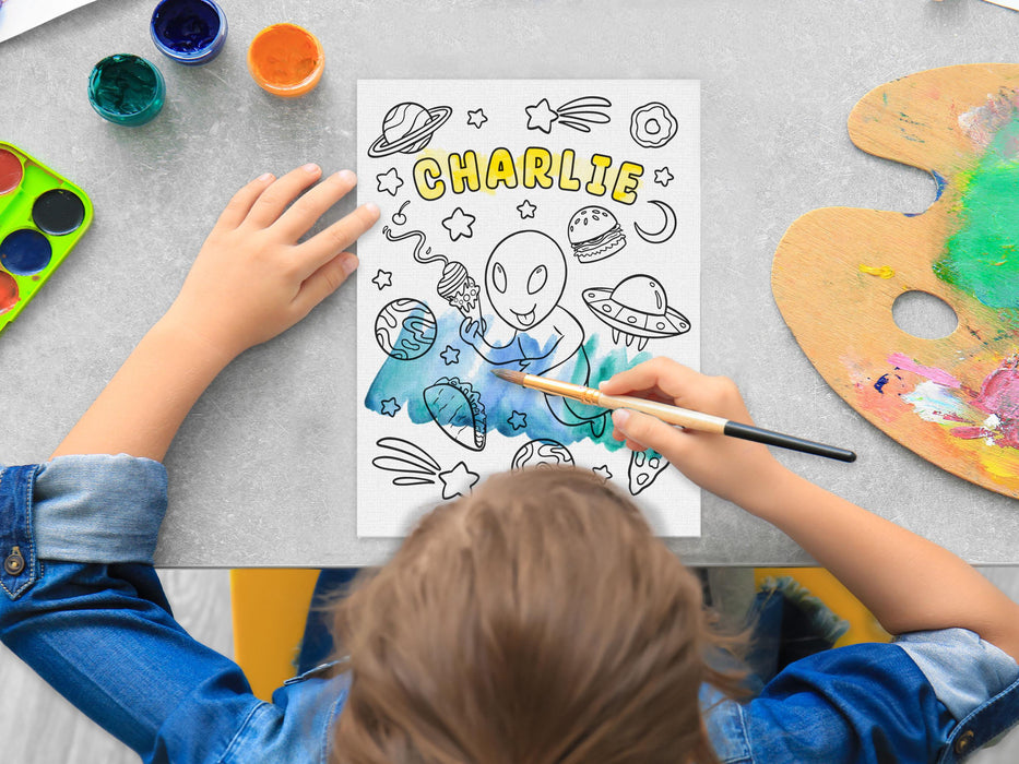 kid painting a canvas panel with a custom space alien adventure design printed on it, kid is using watercolors on grey table surrounded by painting supplies such as cups of paint, palettes, brushes, etc