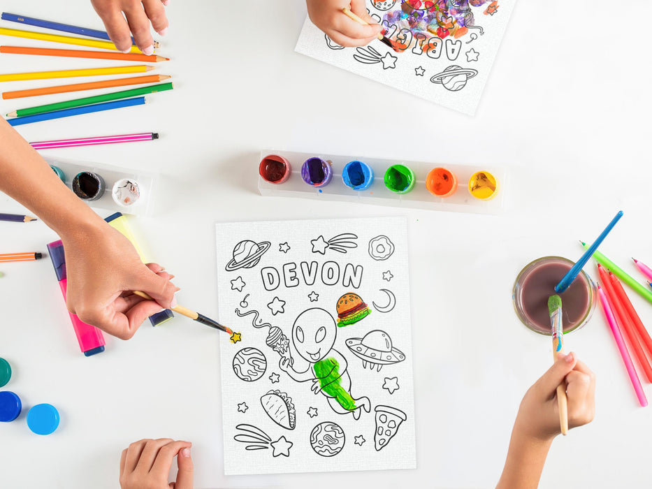 two kids and an adult painting two canvas boards with custom space alien adventure designs printed on it with custom names, placed on white table surrounded by art supplies such as paint, markers, pens, highlighters, etc