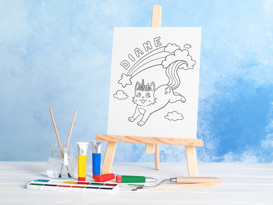 canvas panel with a custom cute caticorn design printed on it sitting on wooden easel, placed on a white wooden table surrounded by painting supplies such as paint brushes, tubes and paint palettes, palette knife, in front of a blue wall