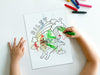 kid at white table coloring a canvas panel with a custom name caticorn design printed on it using crayons with a pile of crayons right next to the canvas