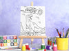 canvas panel with a custom name dinosaur t-rex design printed on it sitting on wooden easel, placed on a white marble table surrounded by painting supplies such as paint brushes, tubes and paint palettes, palette knife, in front of a purple wall