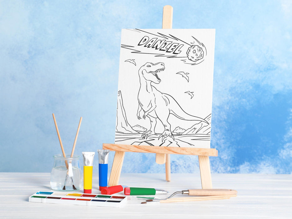 canvas panel with a custom name dinosaur t-rex design printed on it sitting on wooden easel, placed on a white wooden table surrounded by painting supplies such as paint brushes, tubes and paint palettes, palette knife, in front of a blue wall