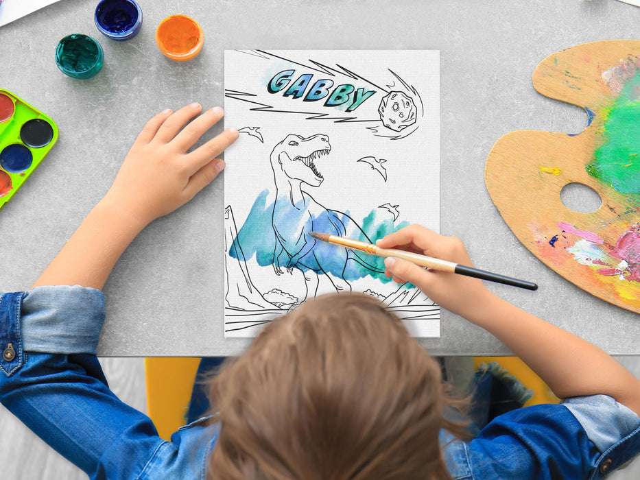 kid painting a canvas panel with a custom name dinosaur t-rex design printed on it, kid is using watercolors on grey table surrounded by painting supplies such as cups of paint, palettes, brushes, etc