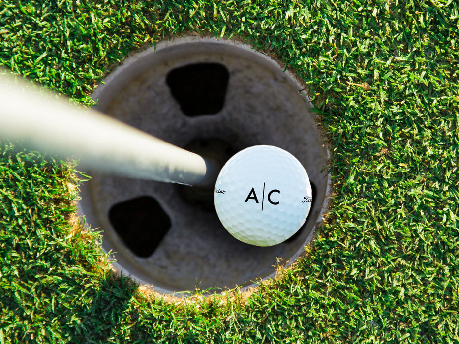 single white titleist golf ball with custom personalized black line monogram printed design in golf course hole next to pole surrounded by golf course grass