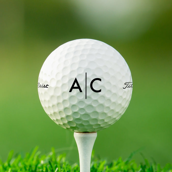 single white titleist golf ball with custom personalized black line monogram printed design on white golf tee against grass golf course background