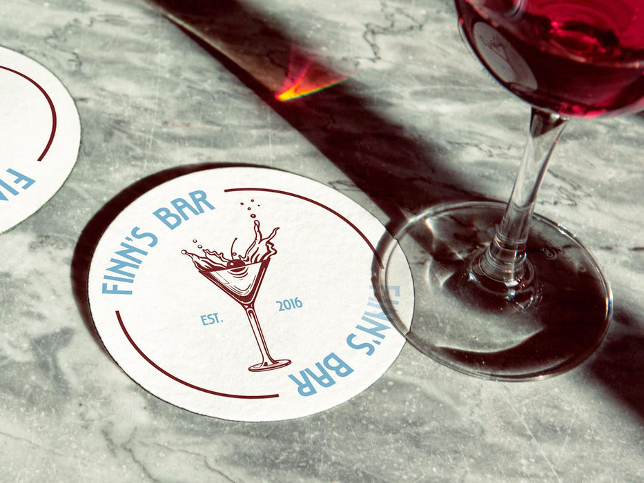 Two coasters sit on marbled surface next to wine glass. Coasters feature bar design. This design uses blue lettering and a drawing of a martini glass. The name, established date, and location can be customized.