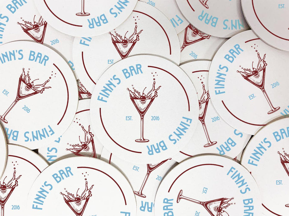 A stack of coasters are spread out across a surface. Coasters feature bar design. This design uses blue lettering and a drawing of a martini glass. The name, established date, and location can be customized.