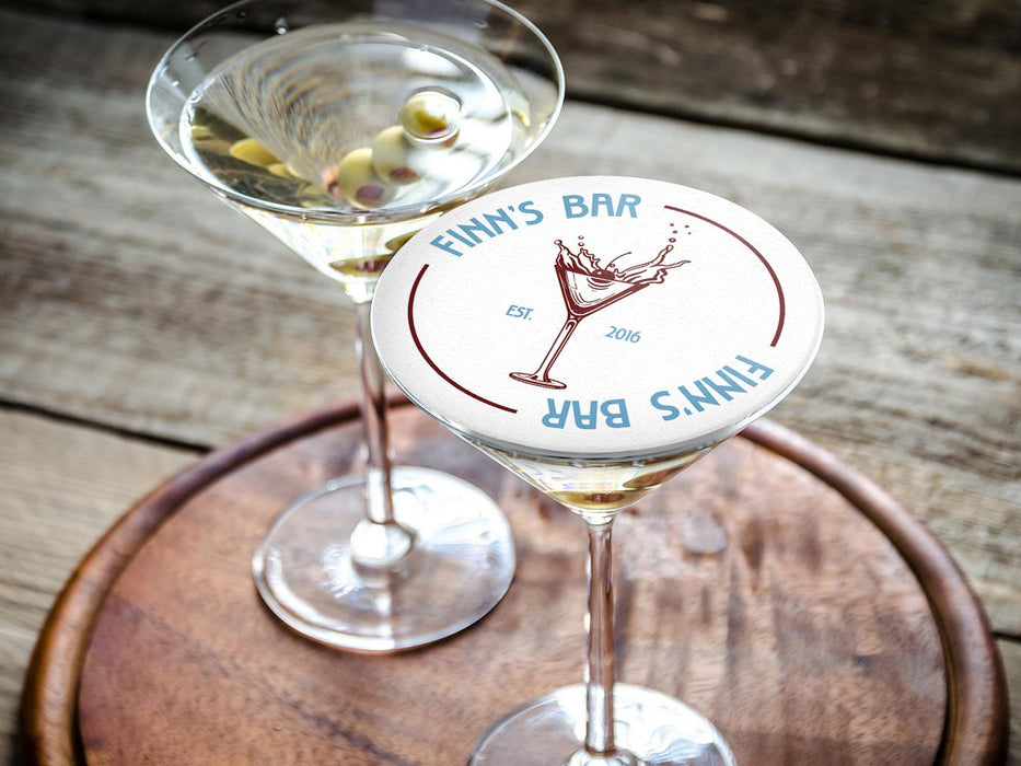 Two martini glasses sits on wooden surface with one glass covered with coaster. Coasters feature bar design. This design uses blue lettering and a drawing of a martini glass. The name, established date, and location can be customized.