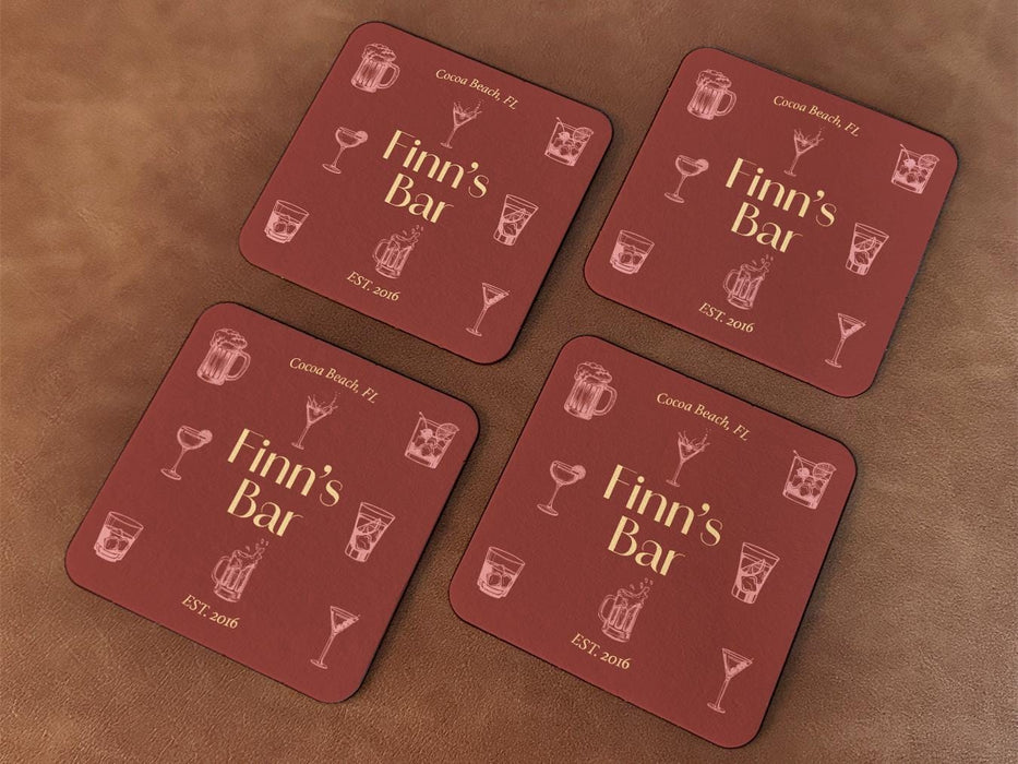 Four coasters sit on a leather background. Coasters feature bar design. This design uses yellow lettering on a red background with drawings of drink glasses. The name, established date, and location can be customized.