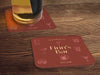 One coaster has a beer glass on it and an empty coaster sits beside it. Coasters feature bar design. This design uses yellow lettering on a red background with drawings of drink glasses. The name, established date, and location can be customized.