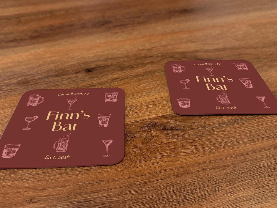 Two coasters sit on a wood background. Coasters feature bar design. This design uses yellow lettering on a red background with drawings of drink glasses. The name, established date, and location can be customized.