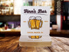 Single coaster sits on top of a stack of coasters. Coasters feature bar design. This design uses brown and orange lettering, and a drawing of beer glasses. The name, established date, and location can be customized.