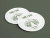 Two coasters sit on dark green surface. Coasters feature bar design. This design uses green lettering on a white background, and a drawing of beer glasses. The name, established date, and location can be customized.