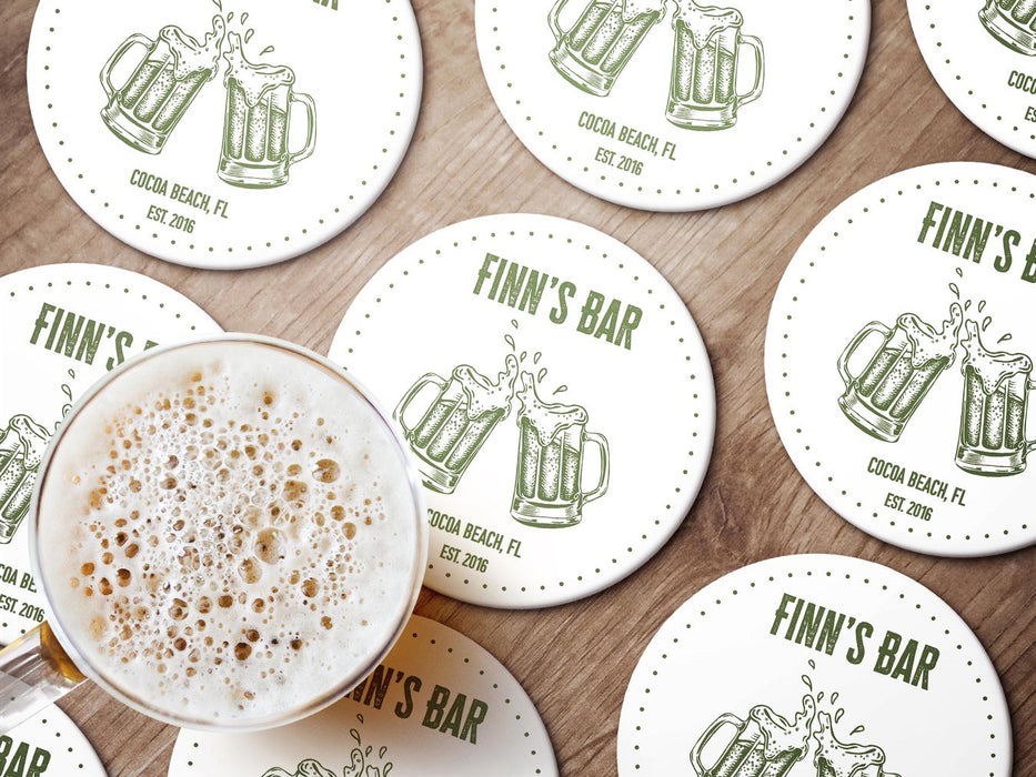 Multiple coasters sit on wooden surface with beer next to them.  Coasters feature bar design. This design uses green lettering on a white background, and a drawing of beer glasses. The name, established date, and location can be customized.