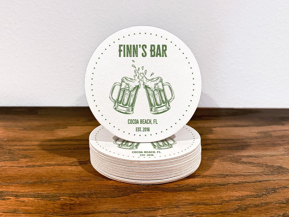 Single coaster sits on top of a stack of coasters. Coasters feature bar design. This design uses green lettering on a white background, and a drawing of beer glasses. The name, established date, and location can be customized.