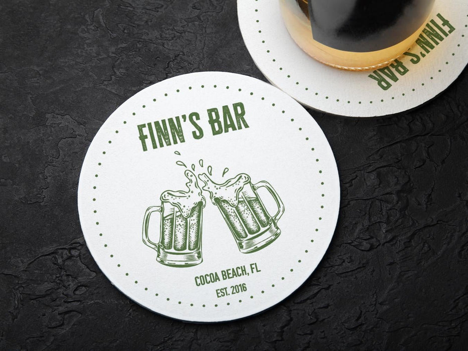 One coaster has a beer glass on it and an empty coaster sits beside it. Coasters feature bar design. This design uses green lettering on a white background, and a drawing of beer glasses. The name, established date, and location can be customized.