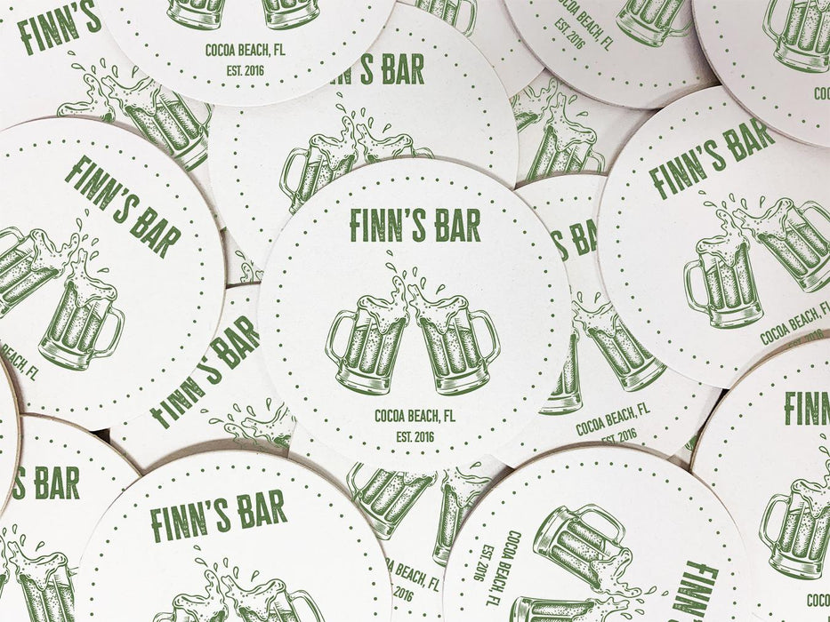 A stack of coasters are spread out across a surface. Coasters feature bar design. This design uses green lettering on a white background, and a drawing of beer glasses. The name, established date, and location can be customized.