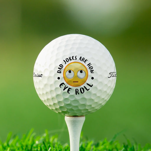 singe white titleist golf ball with dad jokes are how eye roll emoji design on white golf tee in front of golf course grass background 