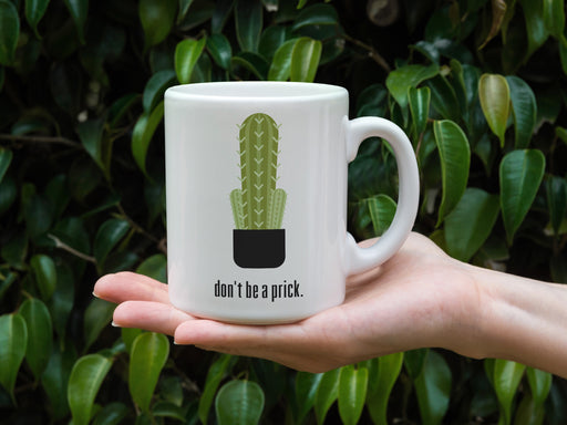 white mug with don't be a prick typography design with cactus graphic being held by a hand in front of numerous outdoor plants