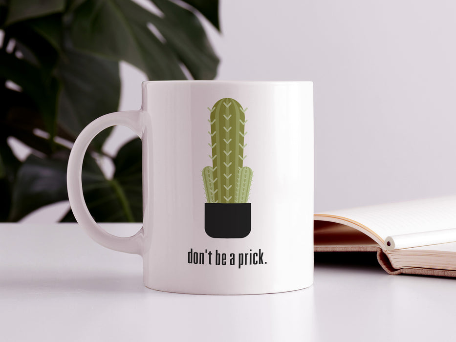 white mug with don't be a prick typography design with cactus graphic on white table next to book in front of house plant