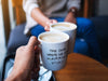 A couple in a cafe holding 2 white mugs filled with coffee together toasting with Friends Themed design typography that says The One Where We Got Married 07.28.23