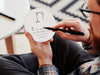 white room with carpet and table with man in jeans and glasses writing on white hangman game coaster with black marker