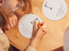 two white hangman game coasters on wooden table with woman writing on single coaster