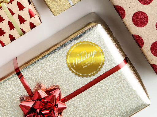 A gold foil cardstock Happy Holidays gift tag is shown on a Christmas present. The present is wrapped in gold paper and has a red bow and ribbon around it. The background of the picture is a white surface.